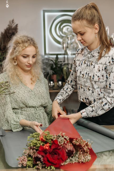 Two women are engaged in floral arrangement within a well-lit workshop or studio. The woman on the right, with long reddish-brown hair tied back, is wearing a patterned blouse and is carefully wrappin
