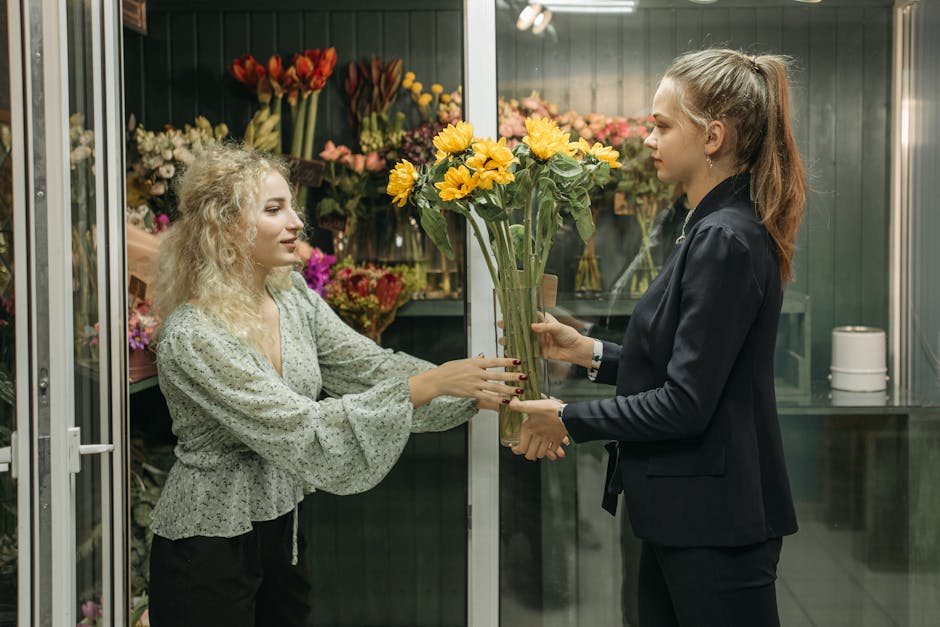 A florist with curly blonde hair, wearing a light green floral blouse, hands a bouquet of bright yellow sunflowers to a customer with long brown hair tied back in a ponytail, dressed in a dark blazer.