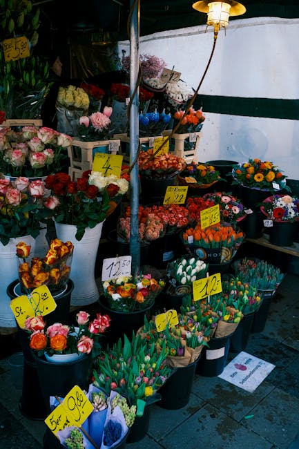 A floral display at a flower market features an assortment of fresh flowers arranged in black and white buckets and baskets. The collection includes pink roses, orange tulips, purple alliums, white hy