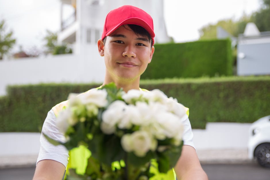 A delivery person wearing a red cap and a high-visibility vest extending a floral gift towards the camera, with a blurred background of residential houses and greenery. The bouquet consists of white f
