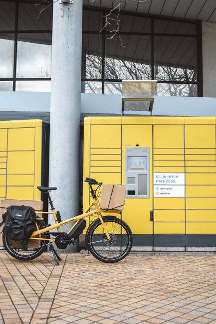 A yellow bicycle with a black seat and a cardboard box attached to the front handlebars is parked in front of a yellow parcel locker at an outdoor location. The parcel locker features multiple compart